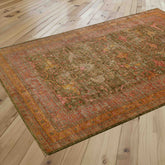 A rectangular vintage-style area rug with an intricate distressed pattern in olive green, rose pink, soft gold, tangerine and crushed raspberry is shown laid out on a light-colored wooden floor.